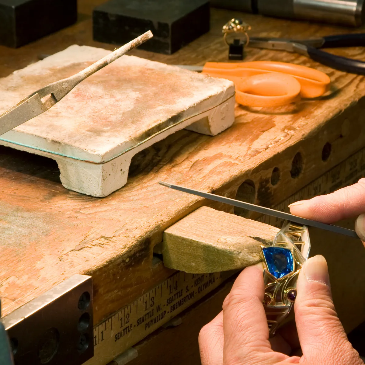 A jeweler resizing a ring with a blue gemstone on a wooden workbench at Masterpiece Jewelers in Daytona Beach.