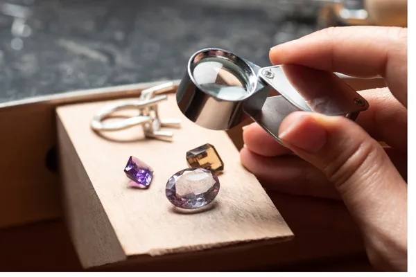 A jeweler inspects gemstones with a magnifying glass, showcasing a ring and various colored stones on a workbench.
