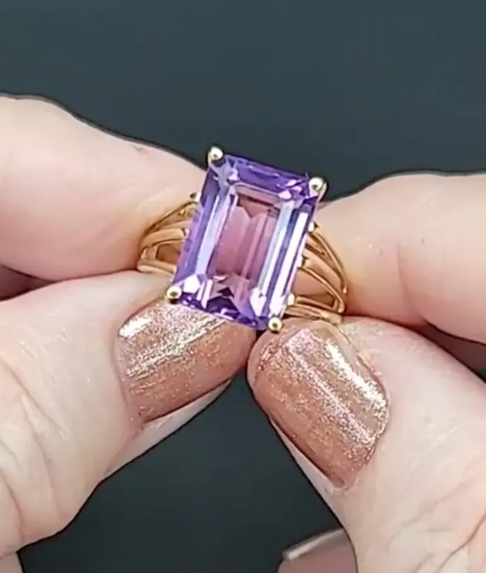 A hand holding a gold ring with a large purple gemstone, showcasing elegant nail polish. Visit Masterpiece Jewelers for more.