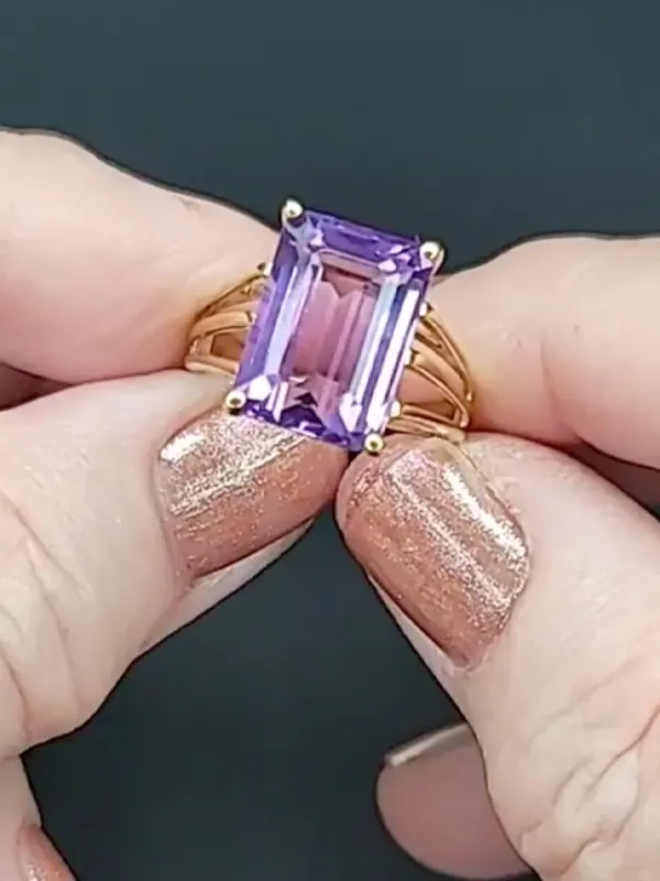 A hand holding a gold ring with a large purple gemstone, showcasing elegant nail polish. Visit Masterpiece Jewelers for more.
