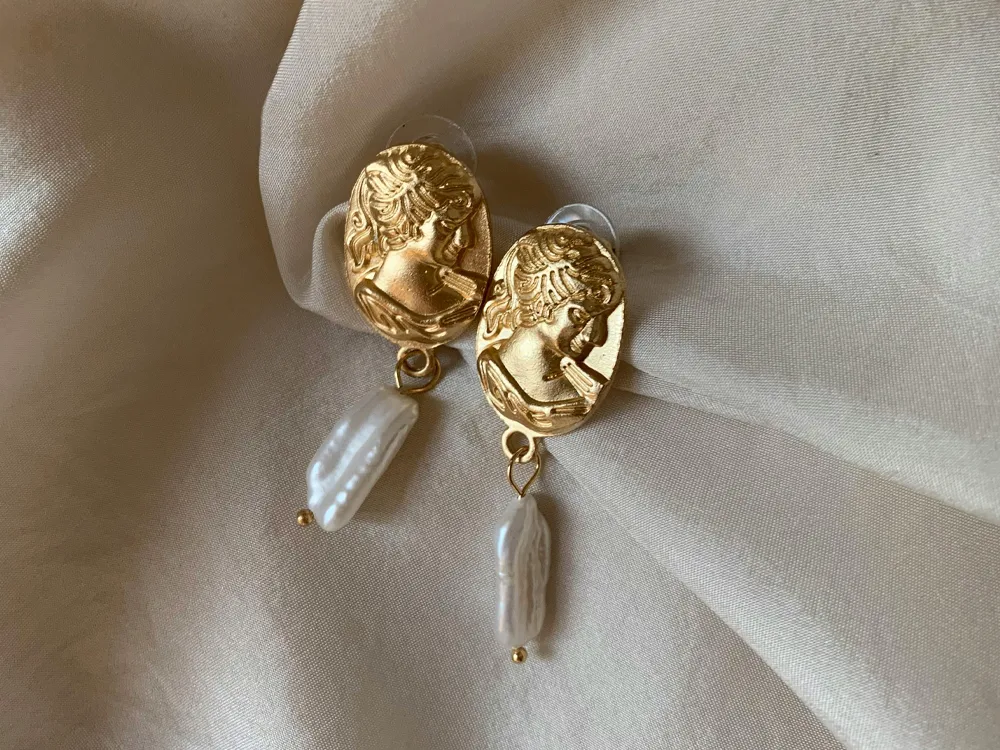 Gold earrings with a vintage design featuring a face and pearl drop. Shop at Masterpiece Jewelers in Daytona Beach.
