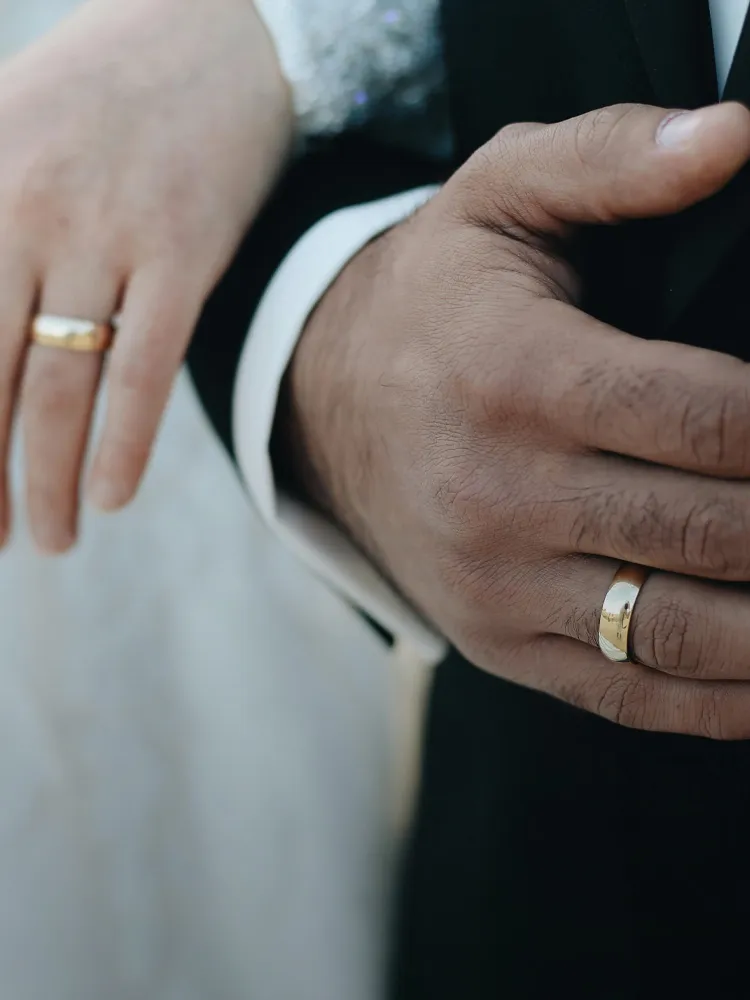 Close-up of a couple's hands wearing wedding bands, symbolizing their union. Visit Masterpiece Jewelers for more.