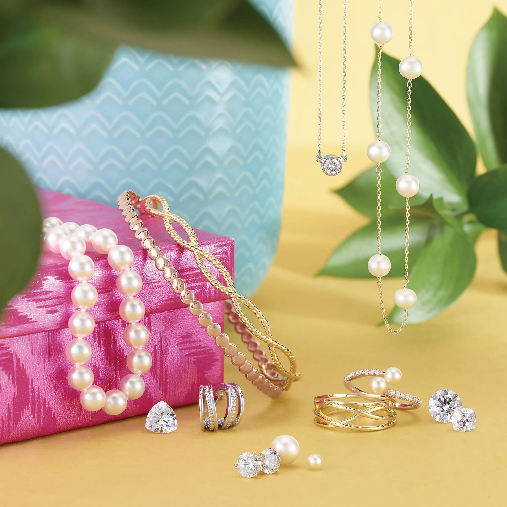 Jewellery displayed on light blue gift box, gold rings, loose diamonds and pearls scattered around with gold necklaces hanging. 