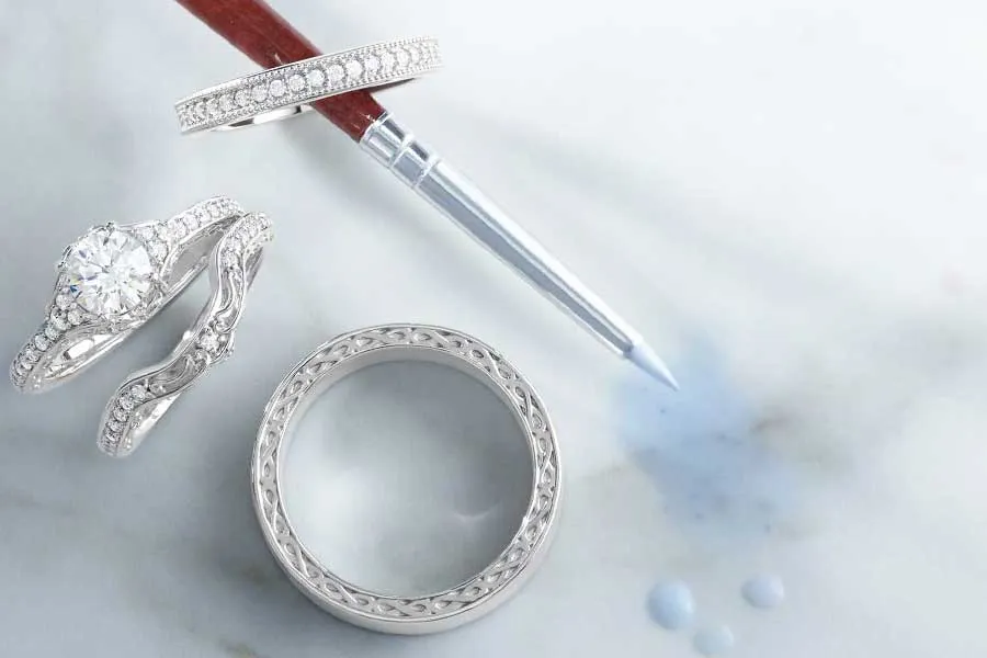 Three elegant white gold rings and a cleaning tool on a marble surface, promoting custom wedding band design at Falls Jewelers. 