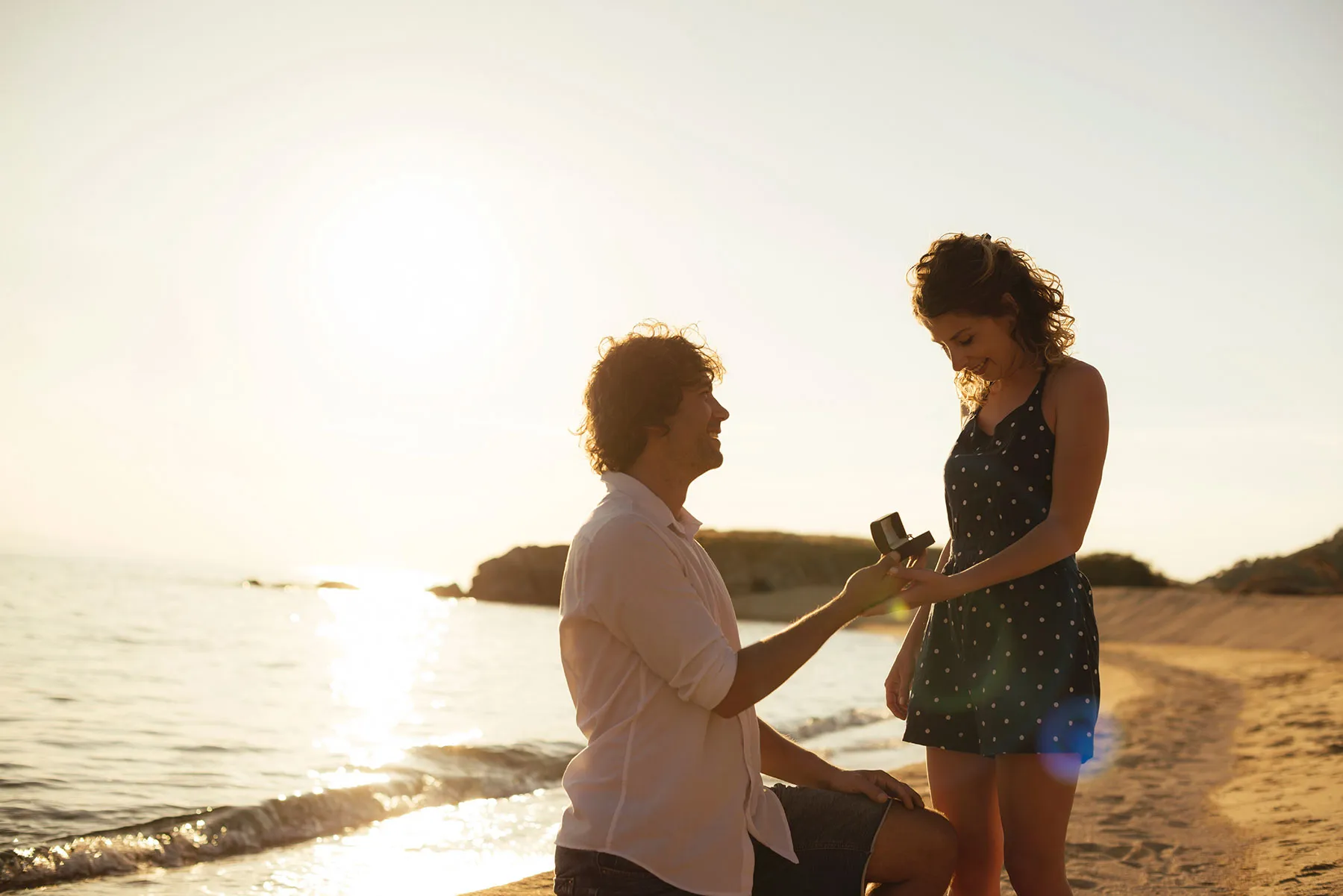 A man proposes to a woman on the beach at sunset, holding an engagement ring in a box.