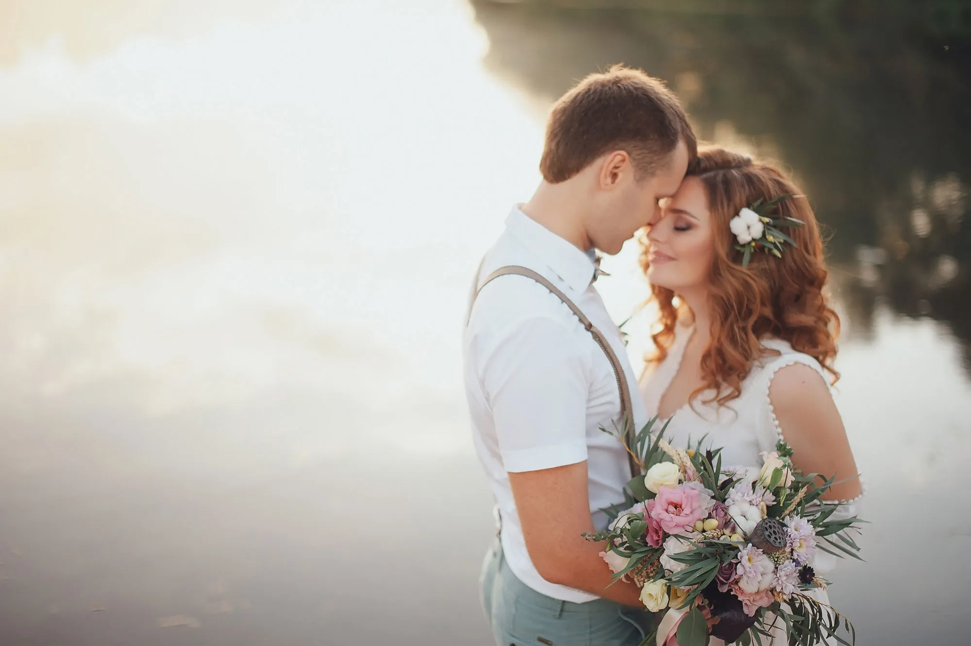 Bride and groom embracing by the water, surrounded by nature, with a bouquet of flowers in the bride's hands. Shop wedding bands