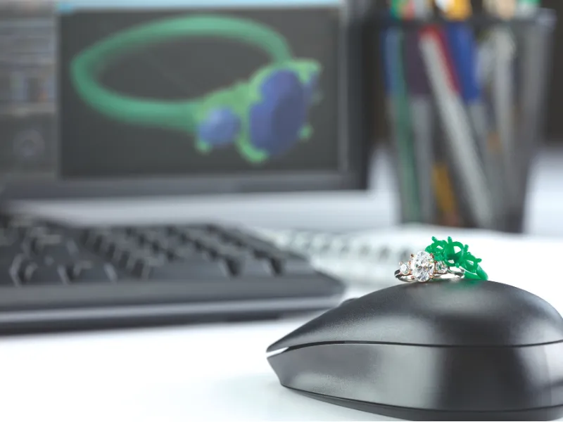 A diamond ring on a computer mouse with a 3D design of a ring displayed on the monitor in a jewelry design setting.