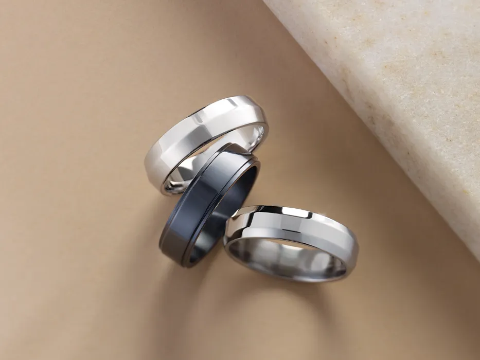 Two silver and one black men's wedding bands on a tan surface