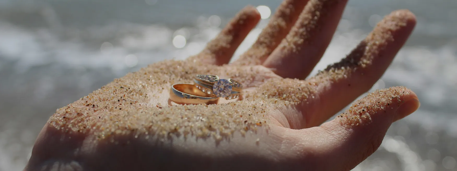 Person holding engagement ring and wedding band in palm with sand all over the palm on a beach