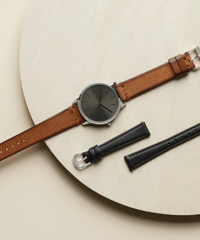 Black watch with brown leather band with black leather band laying next to it on tan background