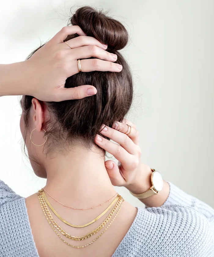 Woman wearing many layered gold necklaces, gold rings, gold hoop earrings, and watch facing away from the camera