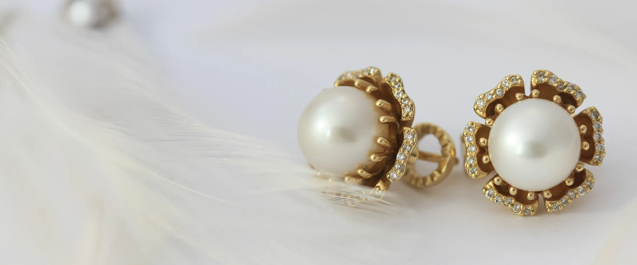 Gold and pearl earrings with diamond accents on a white feather background.