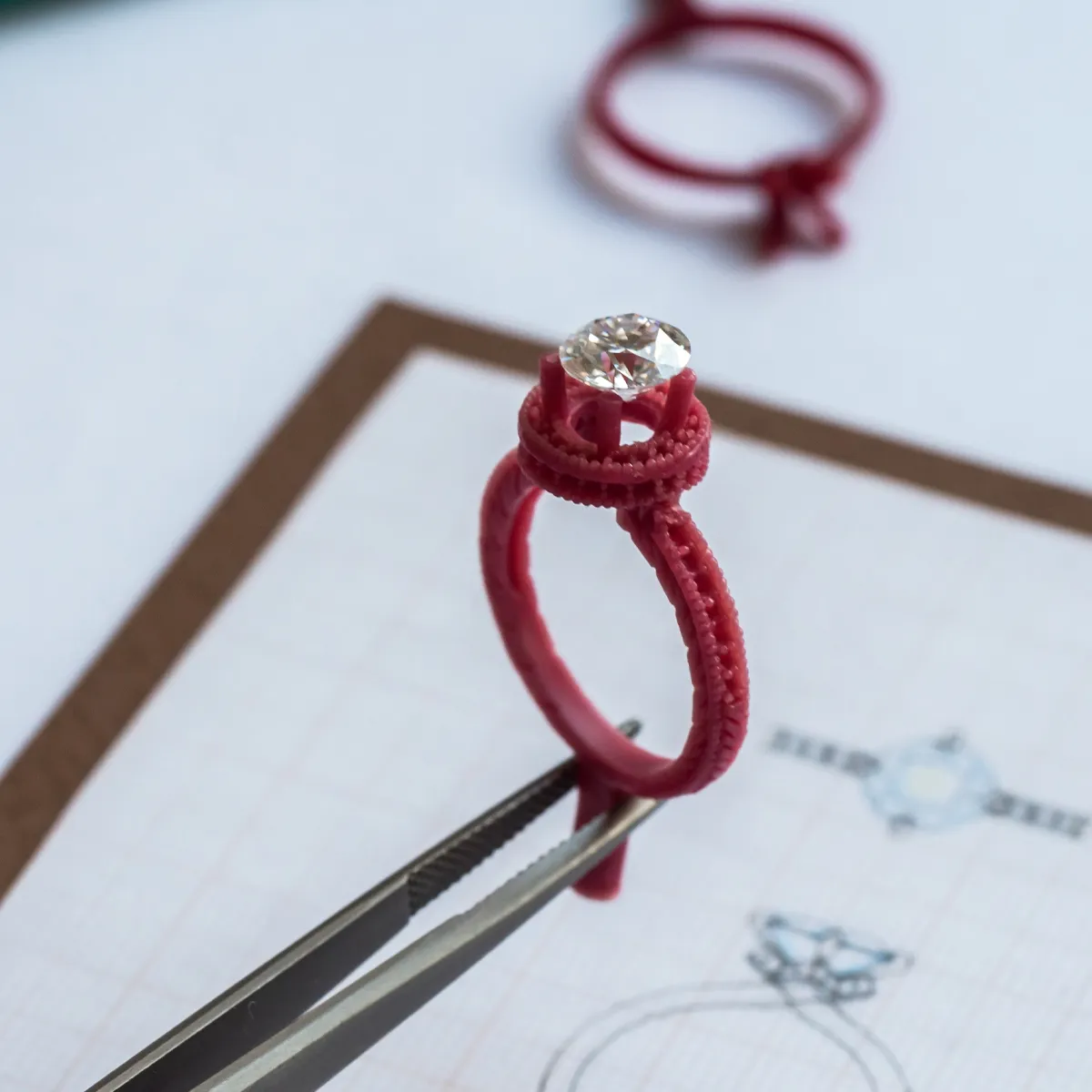 Red wax ring model with a diamond held by tweezers over a design sketch.