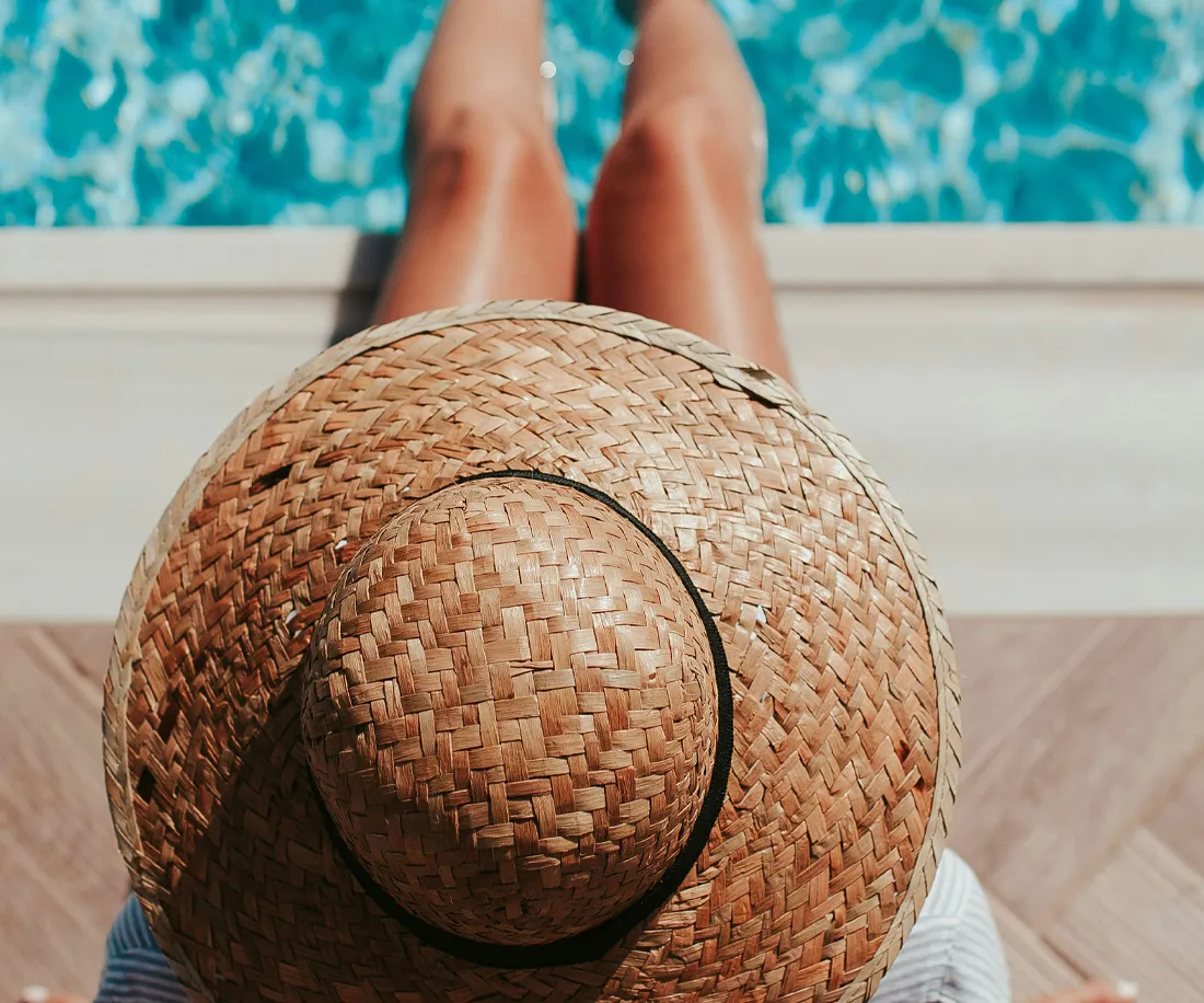 A person relaxing by a pool, wearing a wide-brimmed straw hat, with legs visible in the water.