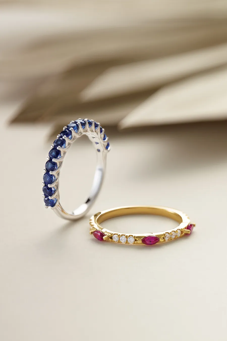 Two elegant rings: one silver with blue stones, one gold with red and clear stones, on a neutral background.