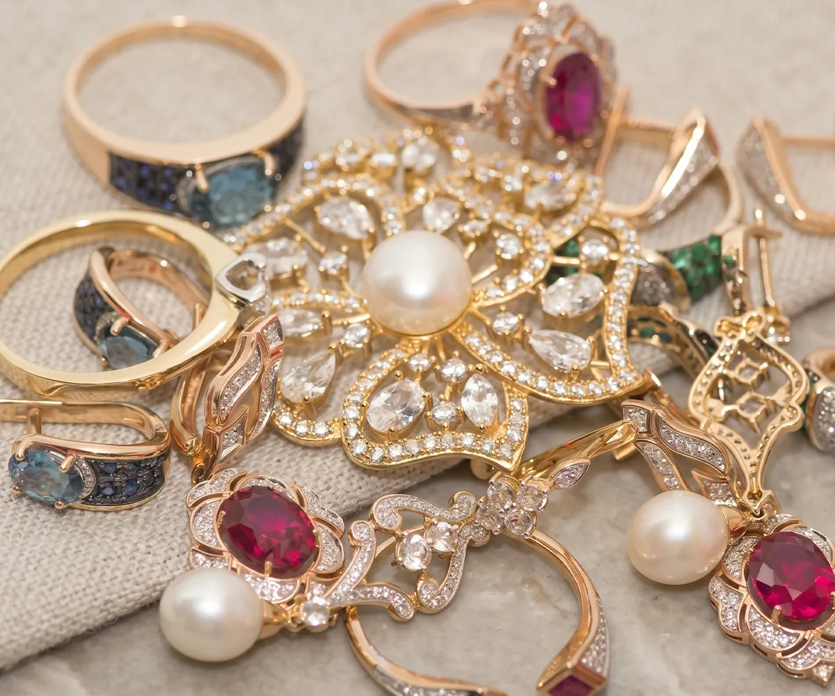 A collection of gold rings and earrings with pearls, rubies, and diamonds on a fabric surface.