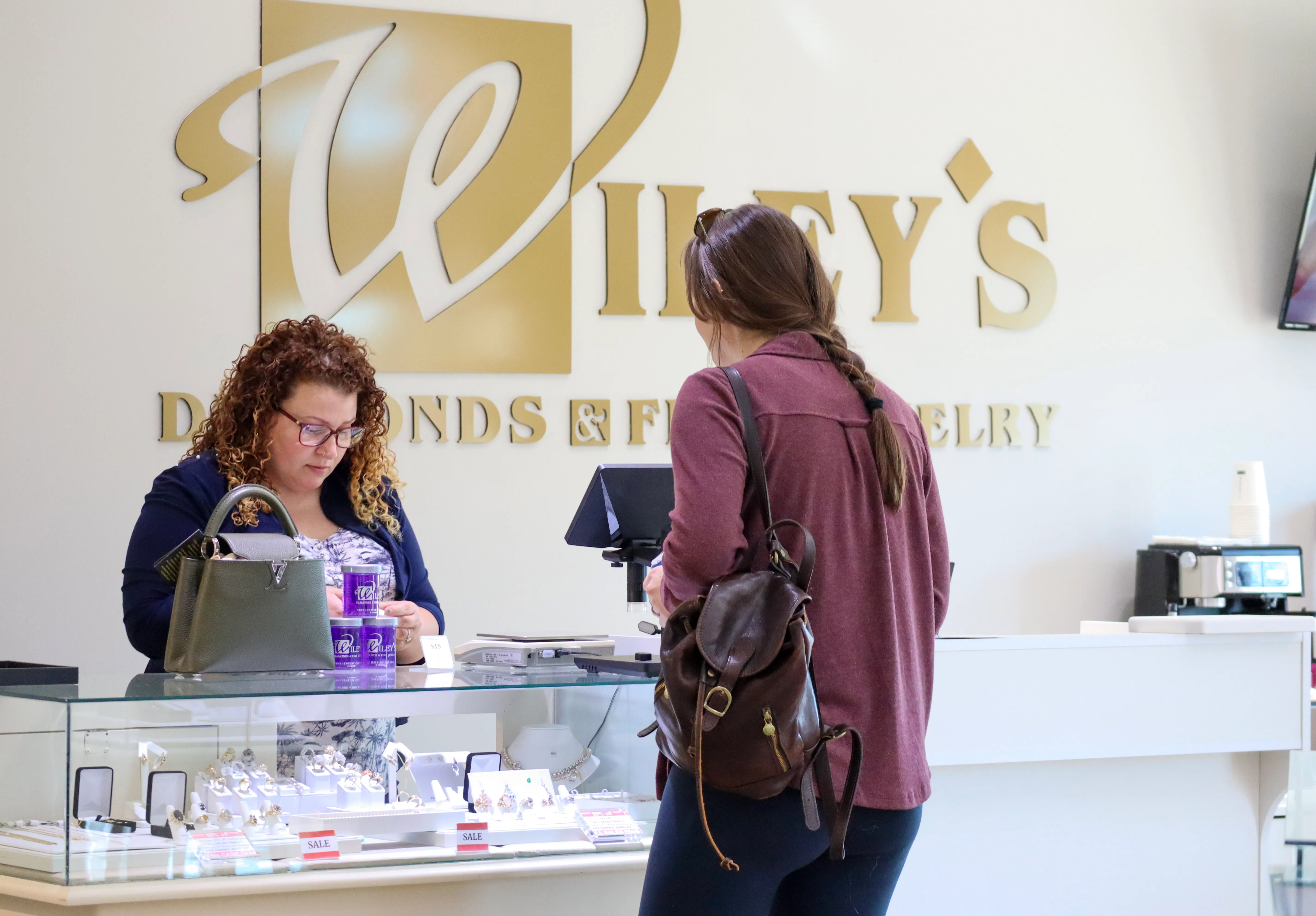 An employee at Wiley's at the counter with a customer as she assesses the customer's jewelry she brought in for repair.