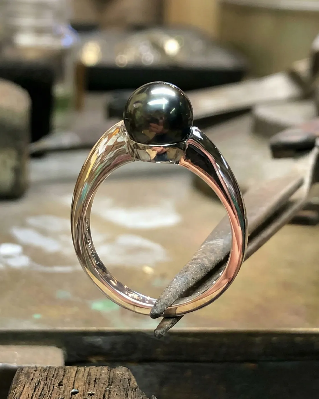 A silver ring with a dark pearl centerpiece is held by a tool in a workshop setting.