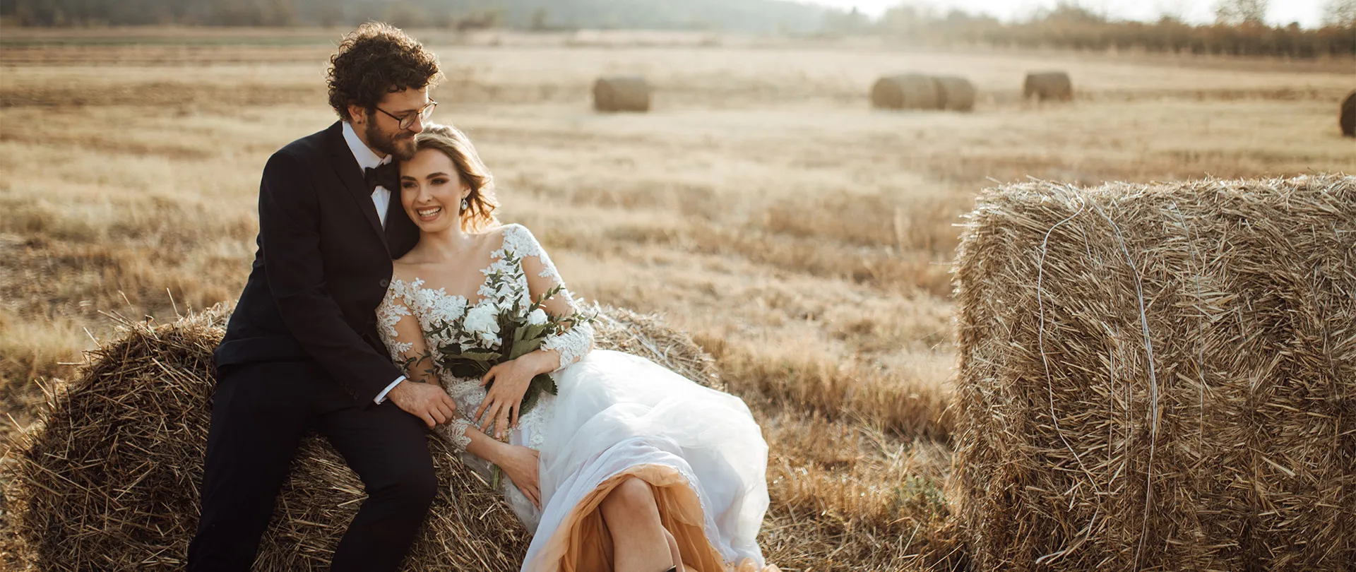 Bride and groom sitting on hay bales in a field, smiling and enjoying their moment together.