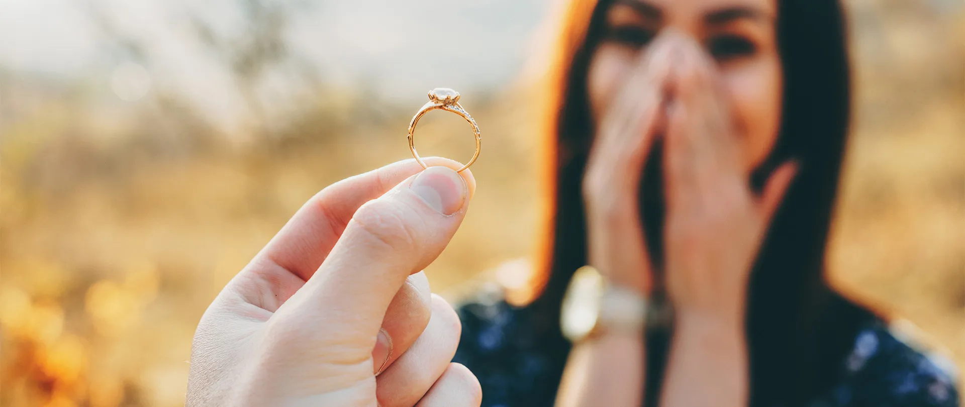 Hand holding an engagement ring with a surprised person in the background. Try Our Ring Builder.