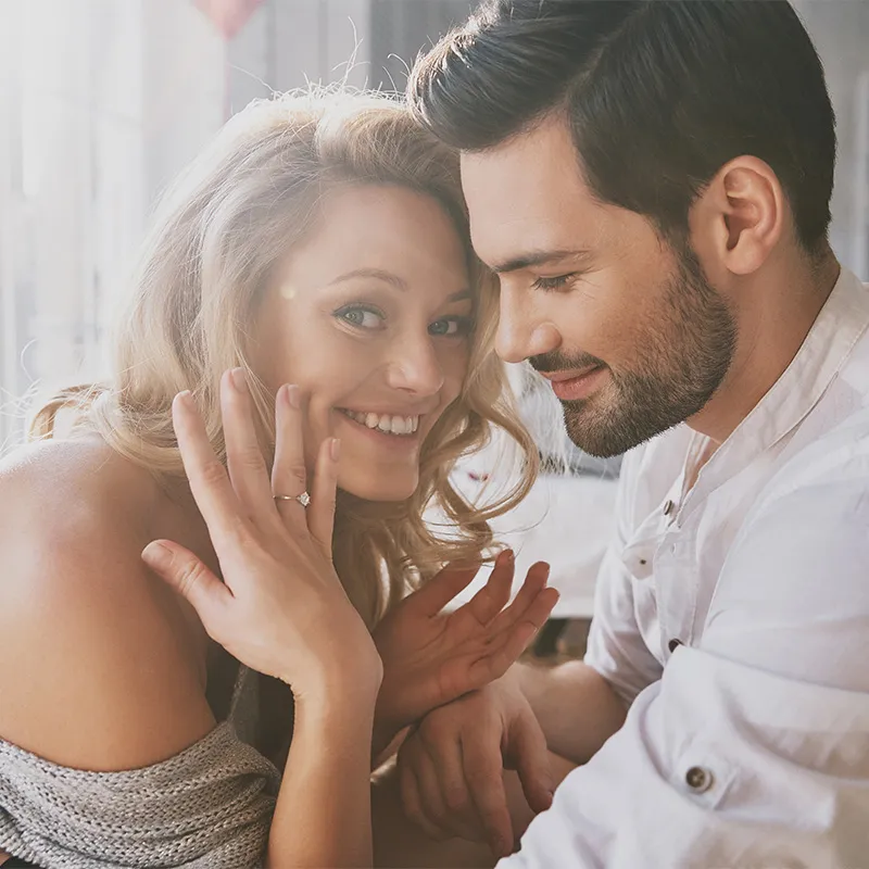 A couple smiling, woman showing engagement ring, in soft sunlight.