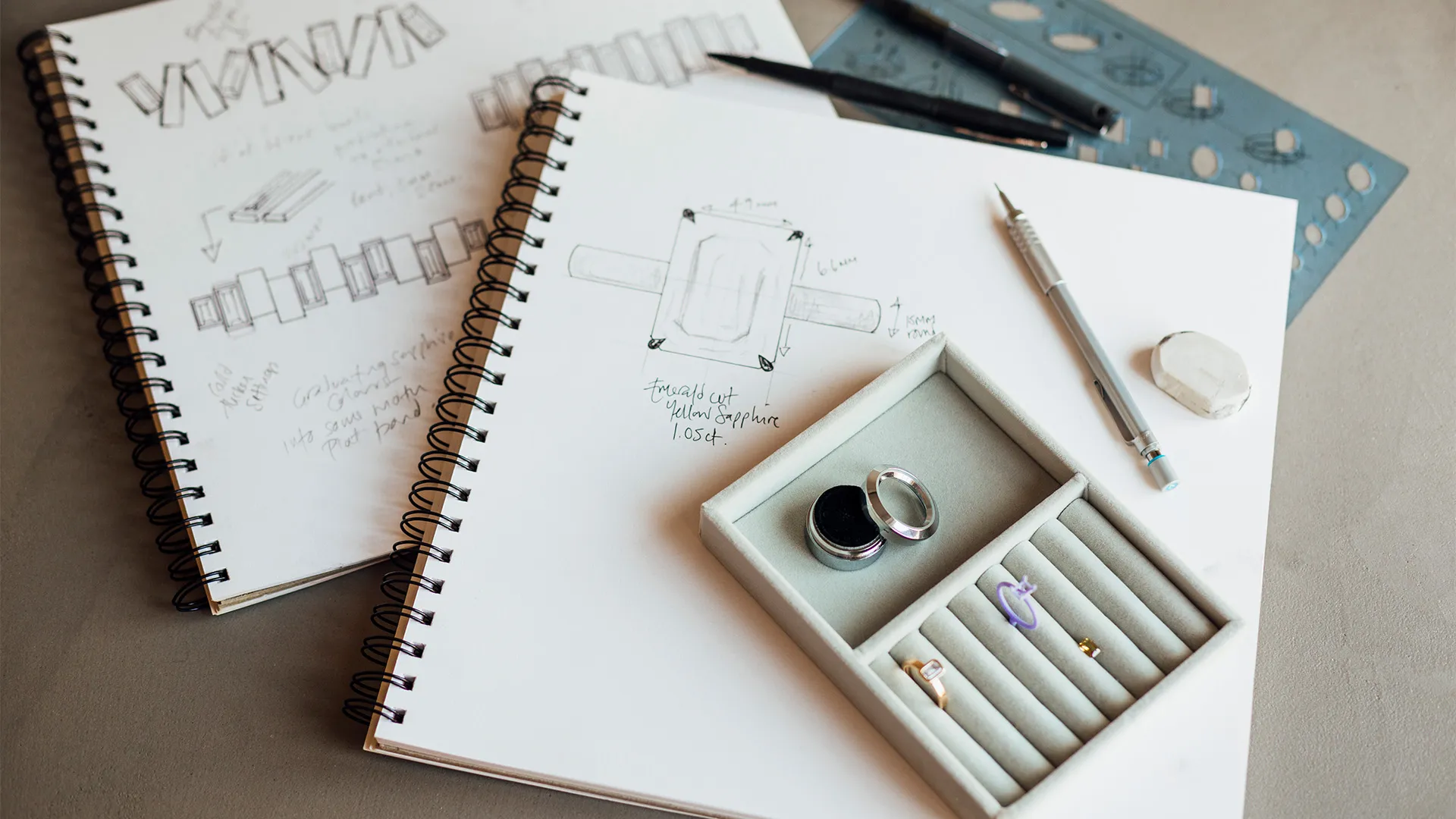 Hands sketching ring designs on paper with jewelry pieces nearby, showcasing creativity in jewelry design at Monarch Jewelry.