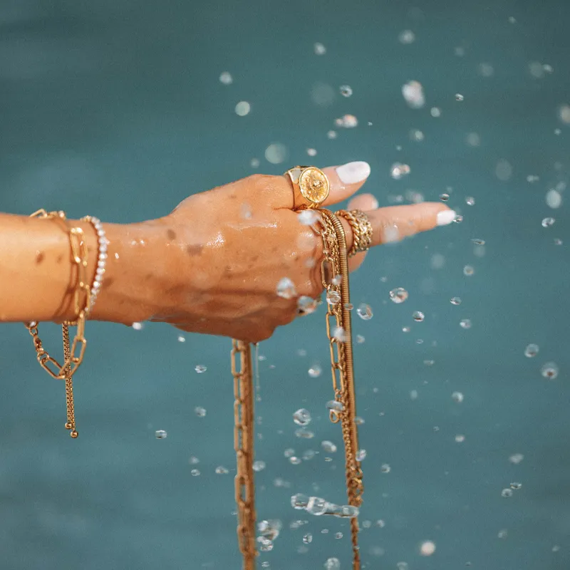 A hand adorned with gold jewelry and rings, holding chains, with water droplets in the air.