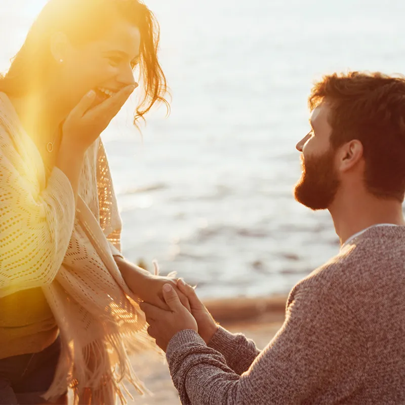 Couple joyfully engaged by the water, capturing a romantic moment at sunset. Visit Quest Fine Jewelers for engagement rings.