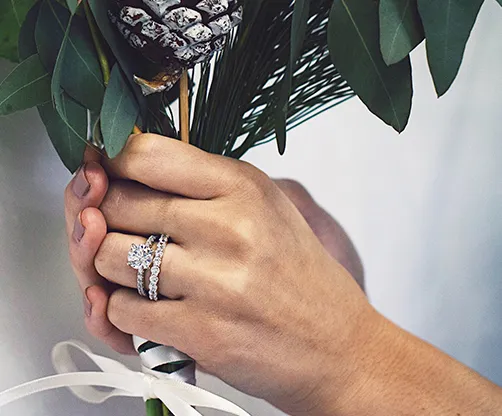 Hand holding a bouquet with wedding rings, promoting Mesa Jewelers' wedding bands. Click to shop wedding bands.